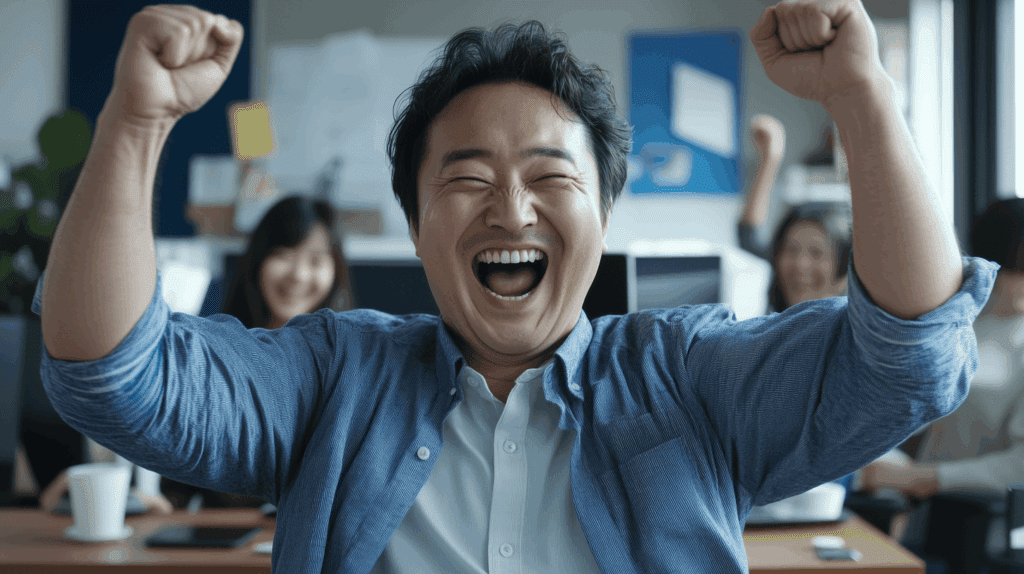 Man celebrating at his desk at the office with coworkers behind him